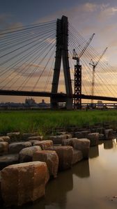 Preview wallpaper bridge, river, rocks, grass, city, building, sky, evening