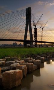 Preview wallpaper bridge, river, rocks, grass, city, building, sky, evening