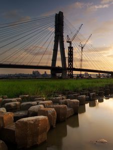 Preview wallpaper bridge, river, rocks, grass, city, building, sky, evening