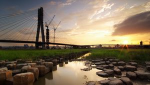 Preview wallpaper bridge, river, rocks, grass, city, building, sky, evening