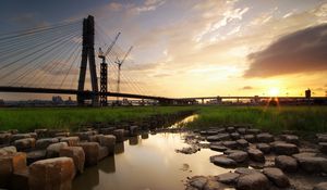 Preview wallpaper bridge, river, rocks, grass, city, building, sky, evening