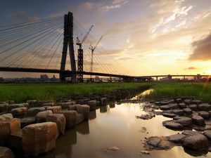 Preview wallpaper bridge, river, rocks, grass, city, building, sky, evening