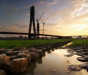 Preview wallpaper bridge, river, rocks, grass, city, building, sky, evening