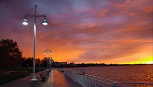 Preview wallpaper city, evening, sunset, sky, clouds, promenade, trees, benches, river, lights