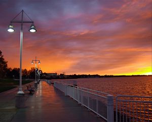 Preview wallpaper city, evening, sunset, sky, clouds, promenade, trees, benches, river, lights