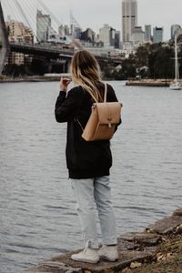 Preview wallpaper girl, backpack, loneliness, sad, shore, river, city