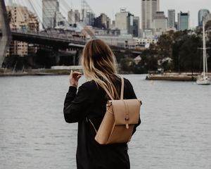 Preview wallpaper girl, backpack, loneliness, sad, shore, river, city