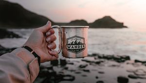 Preview wallpaper mug, hand, water, rocks, sunset